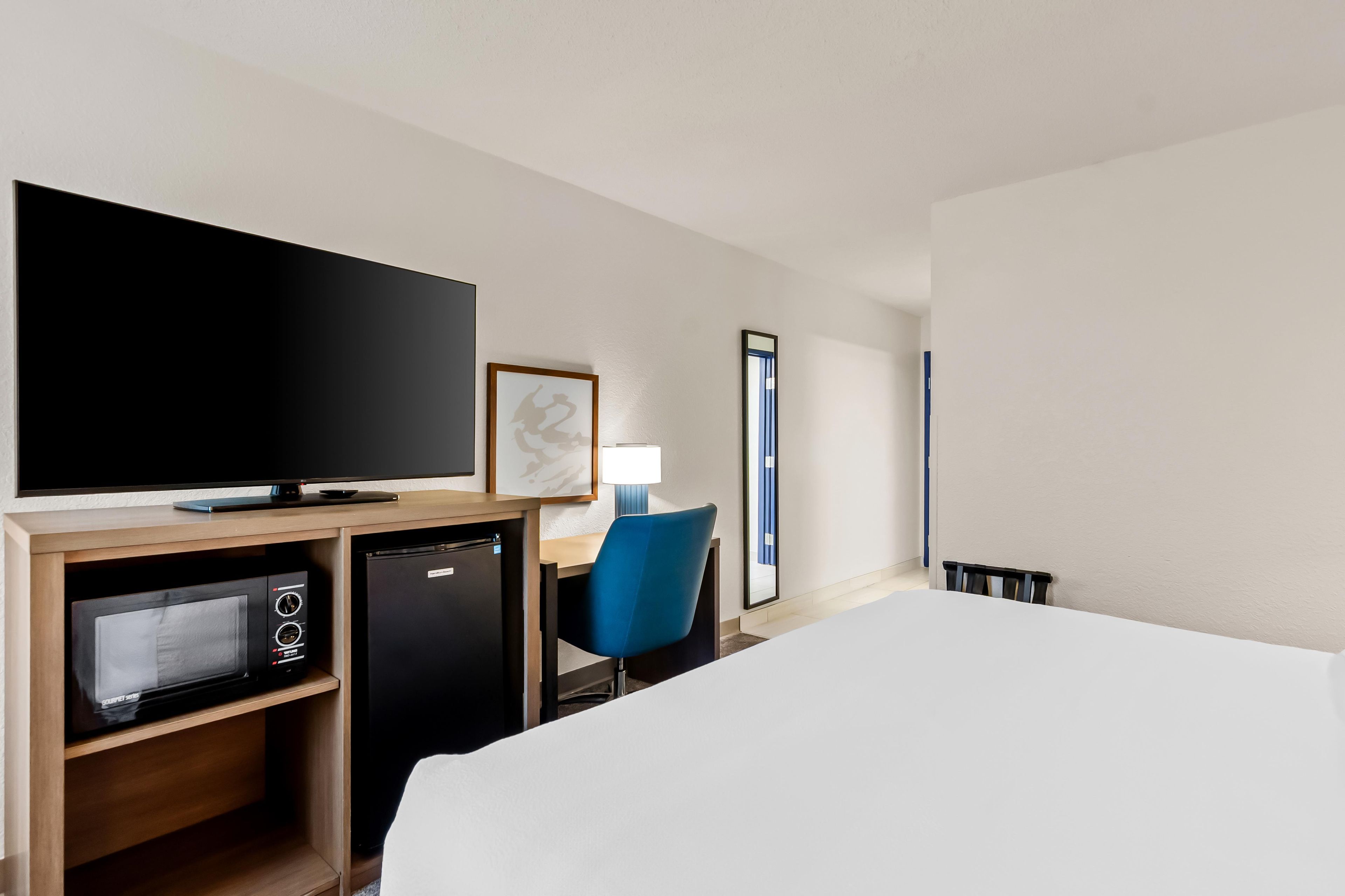 A hotel room with a bed, a television, a desk with a lamp, a chair, and a small refrigerator.