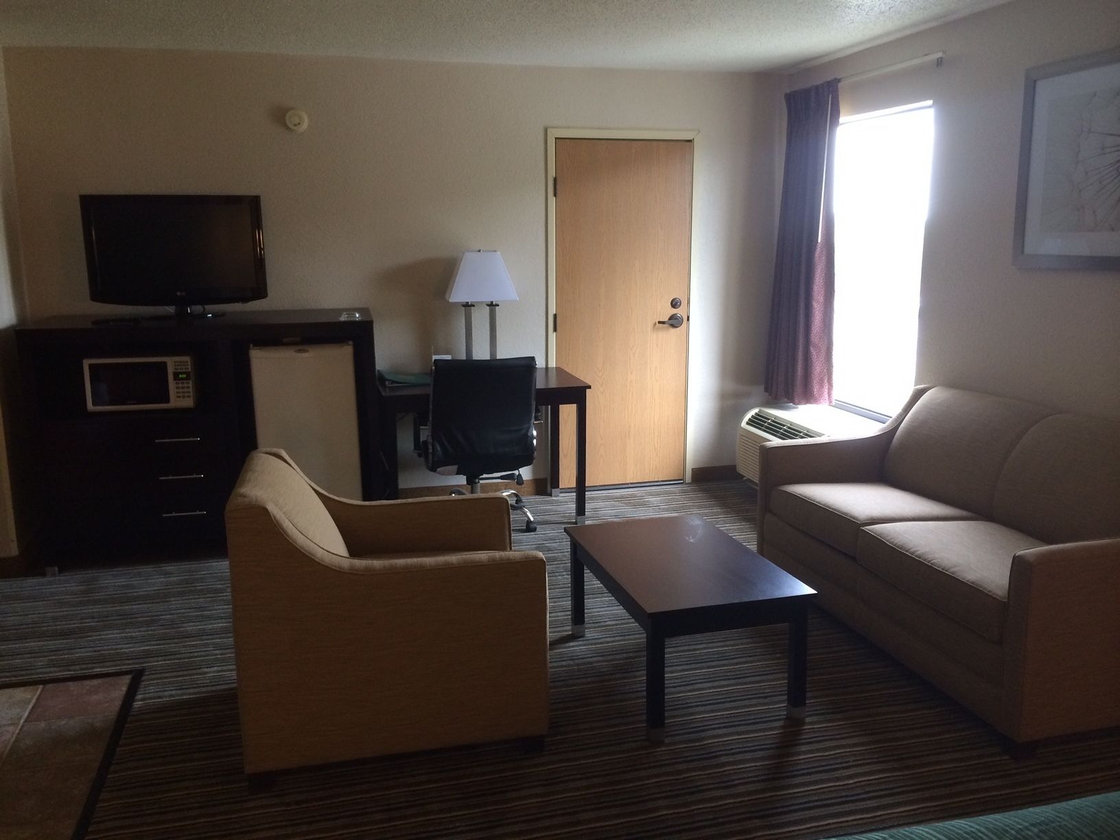 Habitación de hotel con sofá, sillón, televisor, escritorio, lámpara y aire acondicionado.