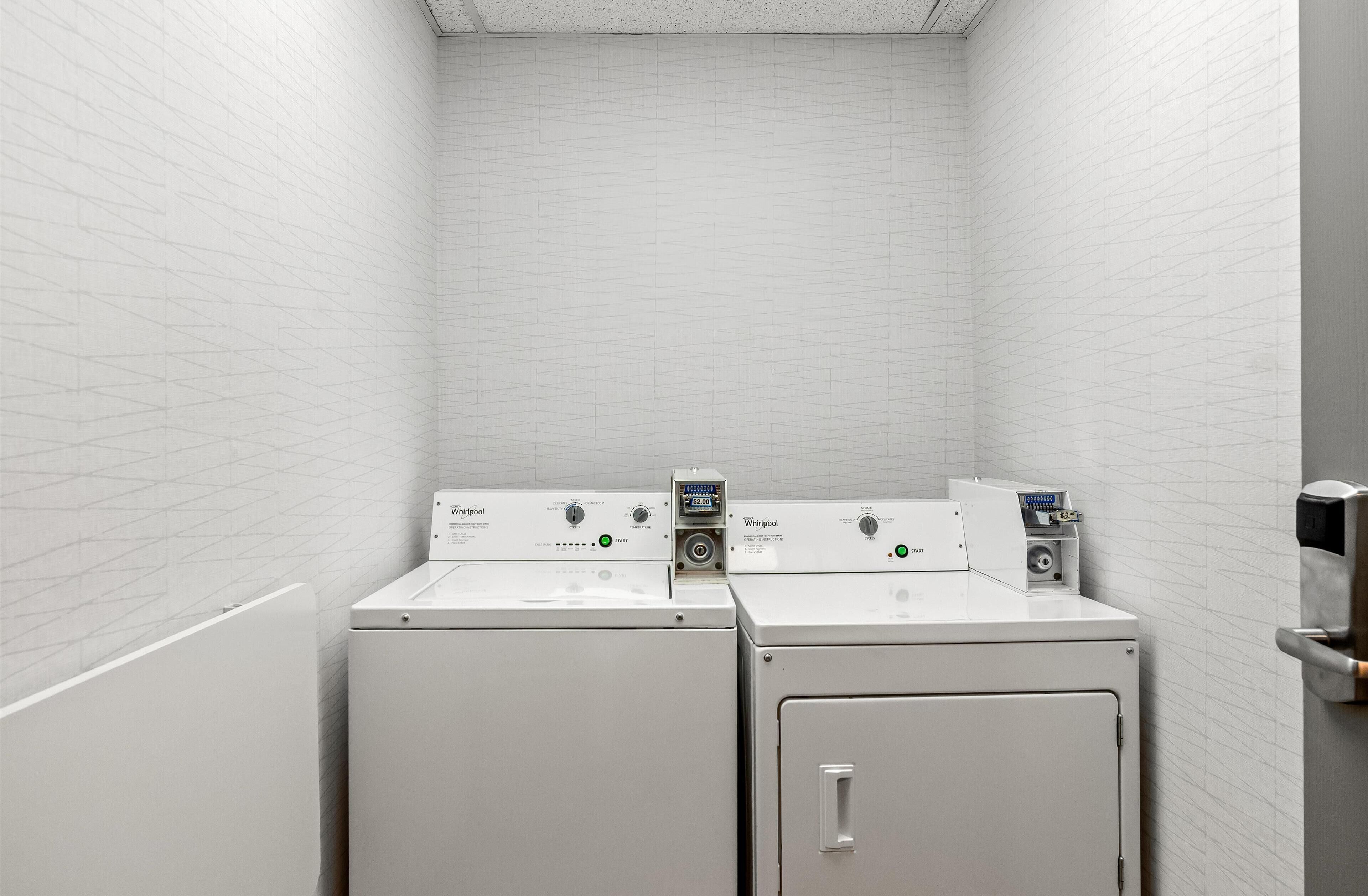 A laundry room with two Whirlpool washers and a dryer, white tiled walls, and a metal door.