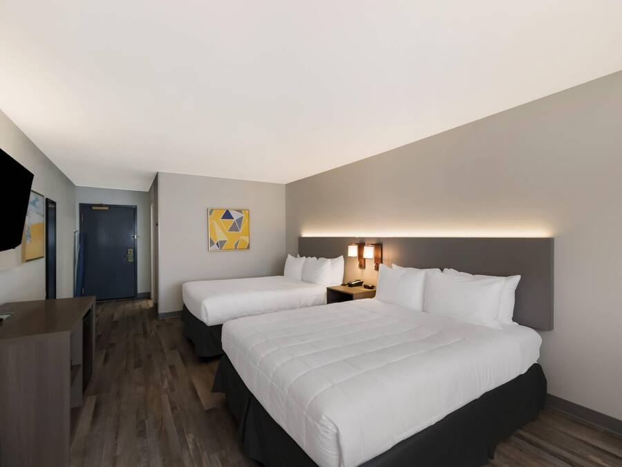Quarto de hotel com duas camas, uma escrivaninha, uma cadeira, uma porta e televisão na parede.