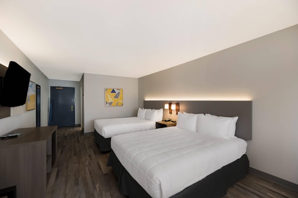 Quarto de hotel com duas camas, uma escrivaninha, uma cadeira, uma porta e televisão na parede.
