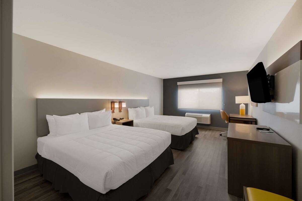 A hotel room with two beds, a desk, a chair, a TV mounted on the wall, and a window with blinds.