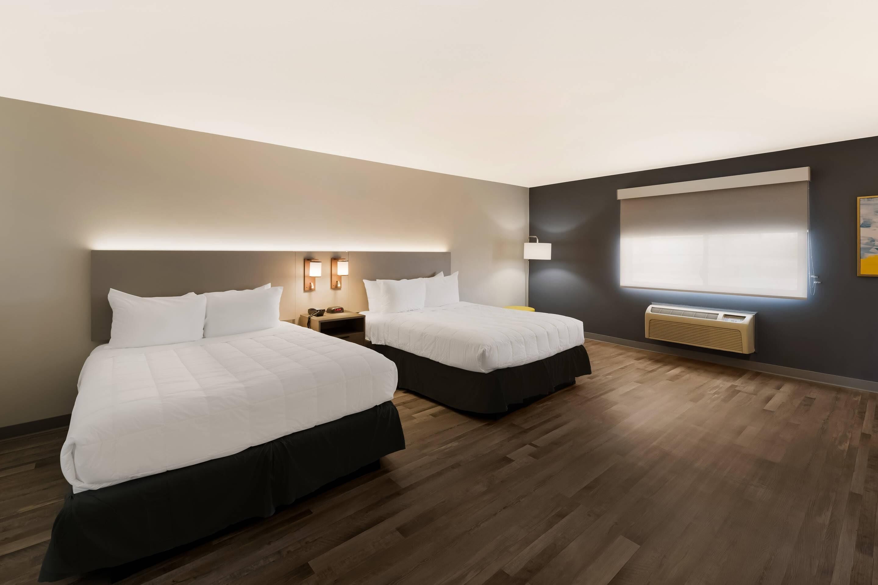A hotel room with two beds, a nightstand, a lamp, a window with blinds, and a radiator.