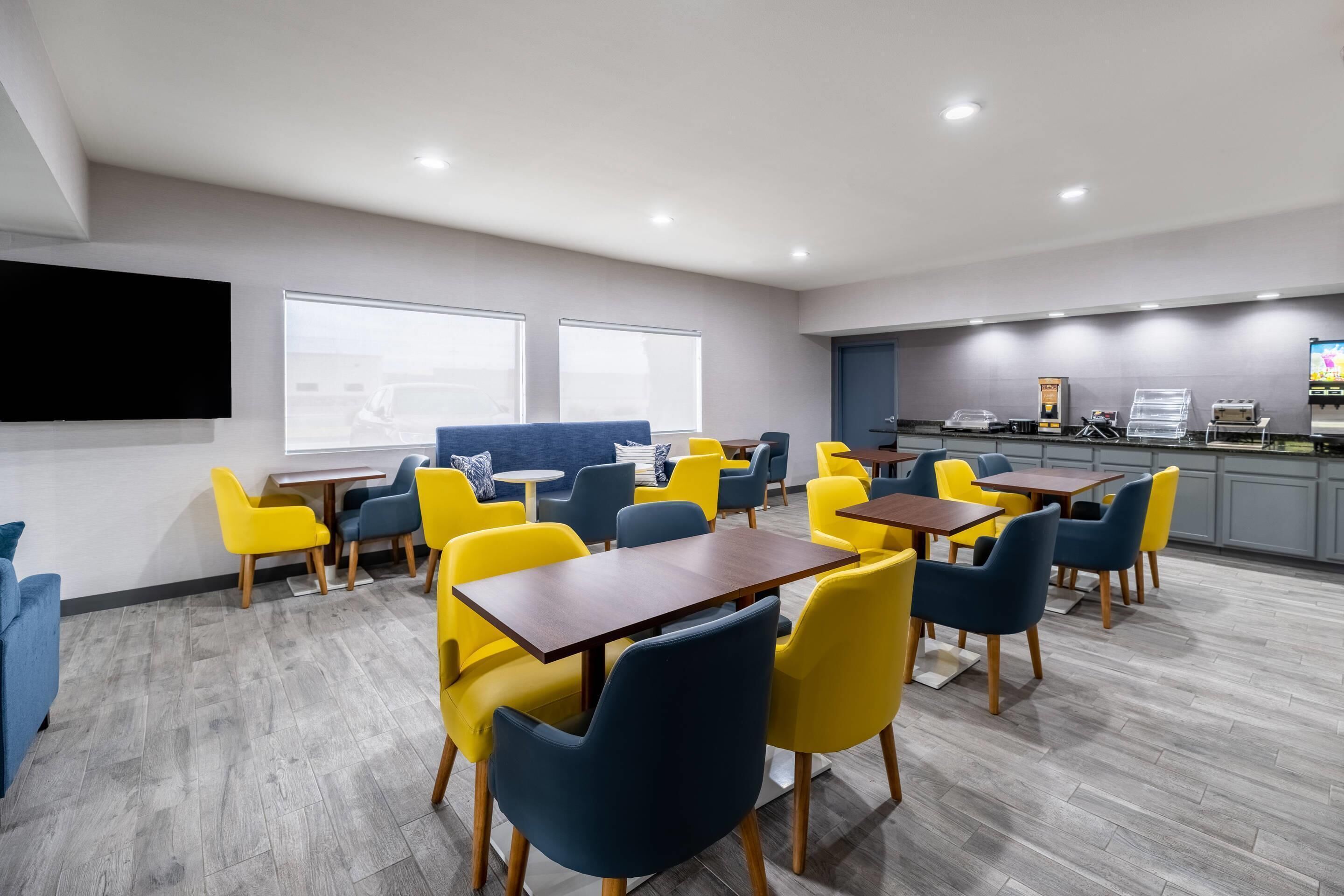 Une salle de conférence moderne, équipée de tables et de chaises, d’un grand écran et d’un coin cuisine avec des appareils électroménagers.