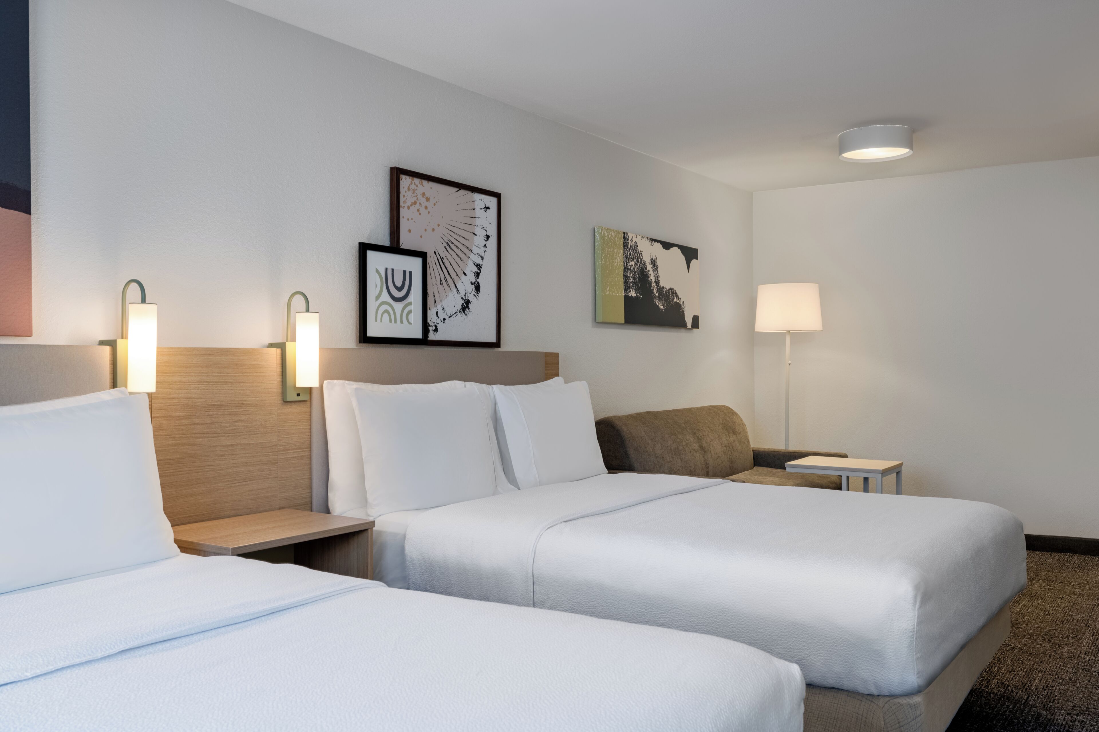 A hotel room with two beds, a nightstand, a lamp, and two wall-mounted artworks.