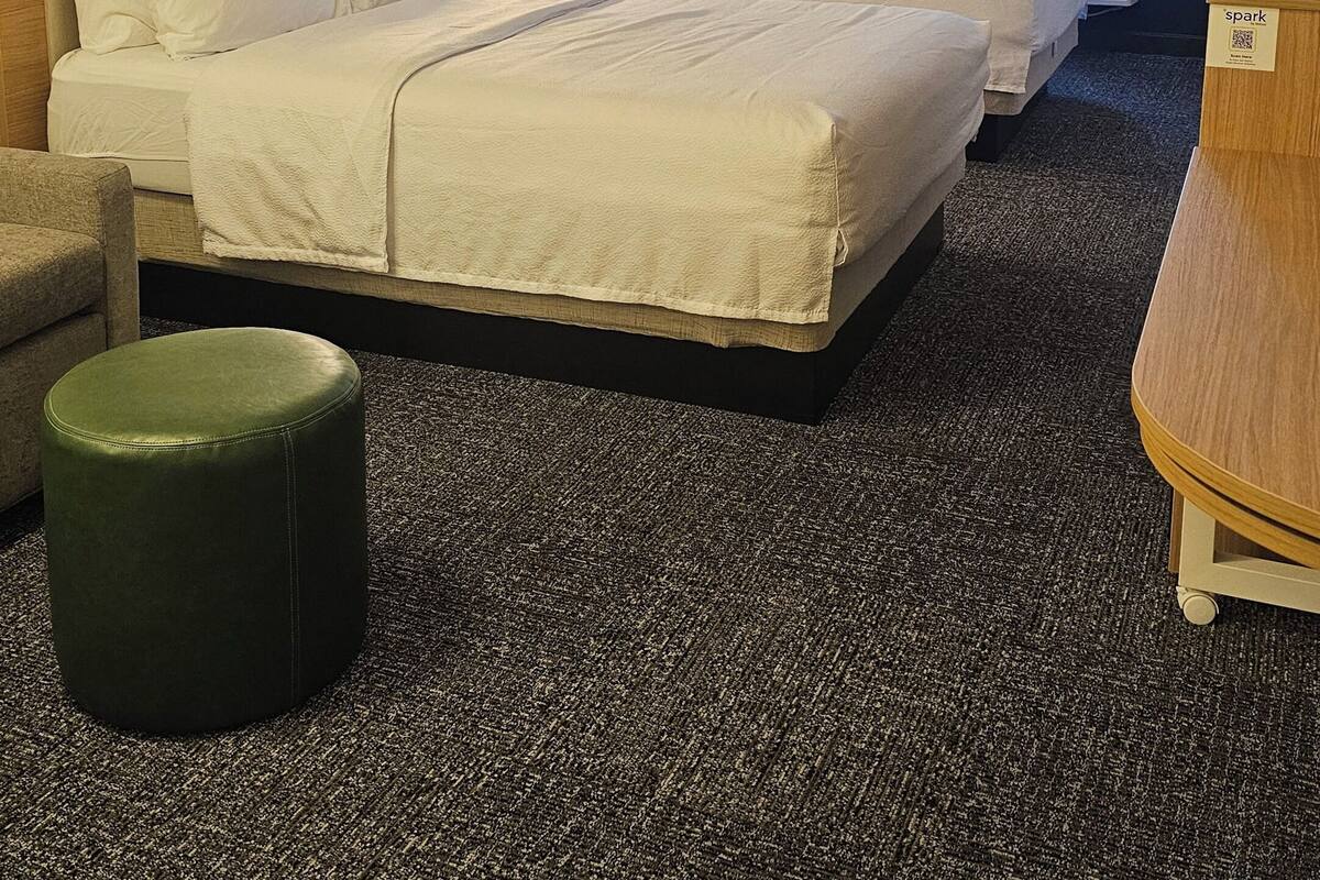 A hotel room with two beds, a green ottoman, a desk, and a window with curtains.