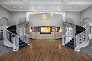 Un vestíbulo de hotel con un mostrador de recepción central, dos escaleras de caracol y una gran obra de arte en la pared.