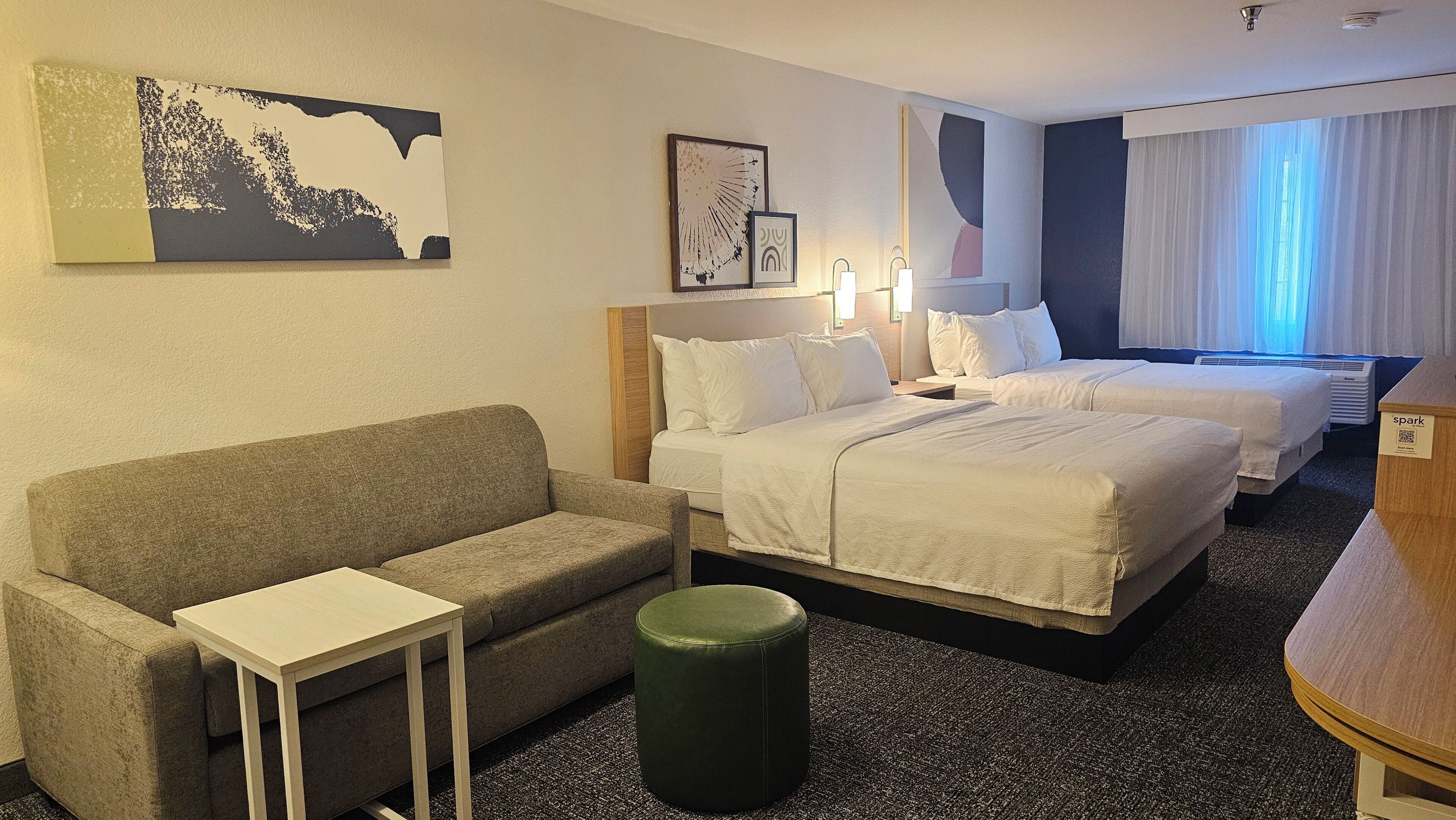 A hotel room with a sofa, two beds, a small table, and a green ottoman.