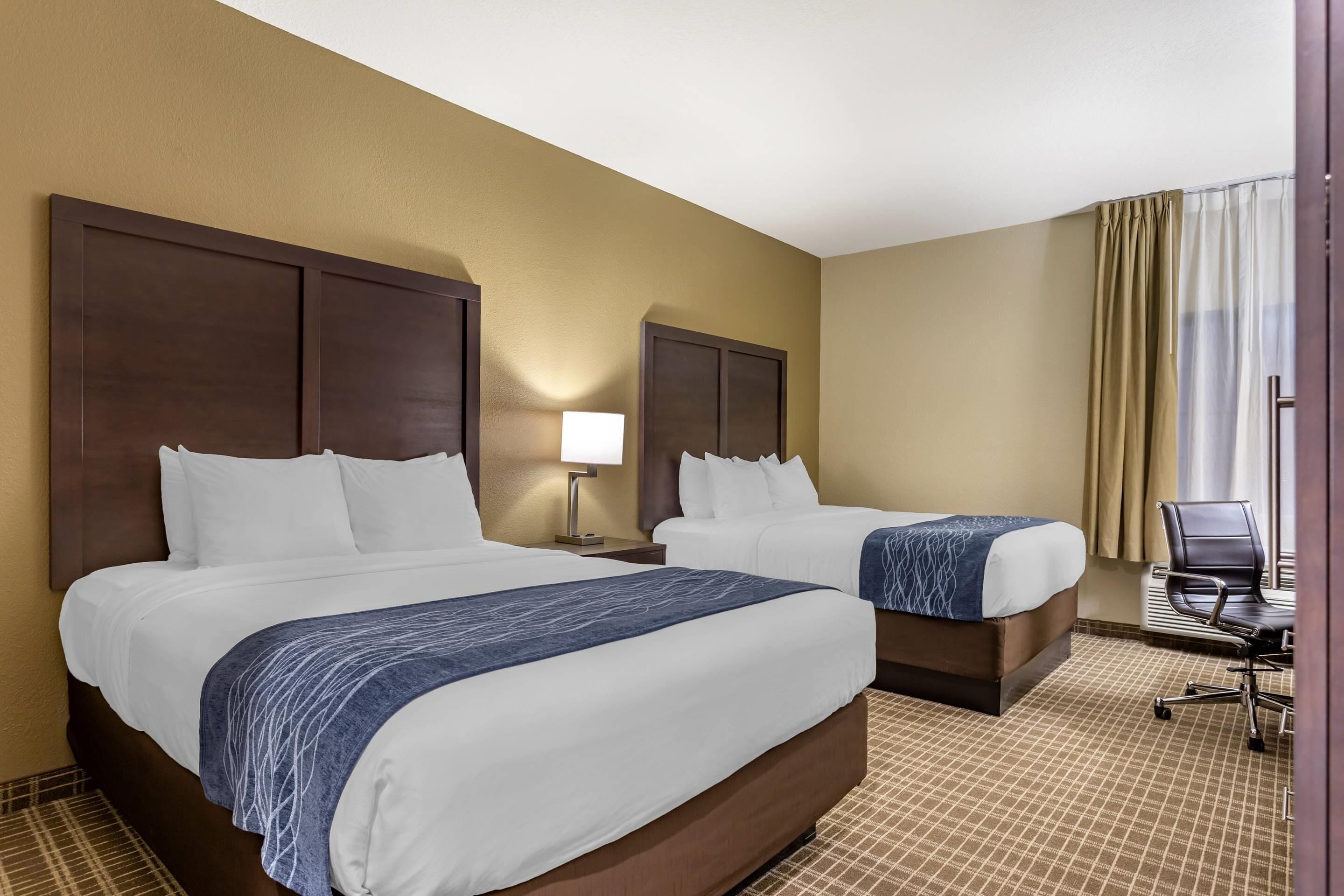 A hotel room with two beds, a nightstand with a lamp, a chair, and a view of the outside through a window with curtains.