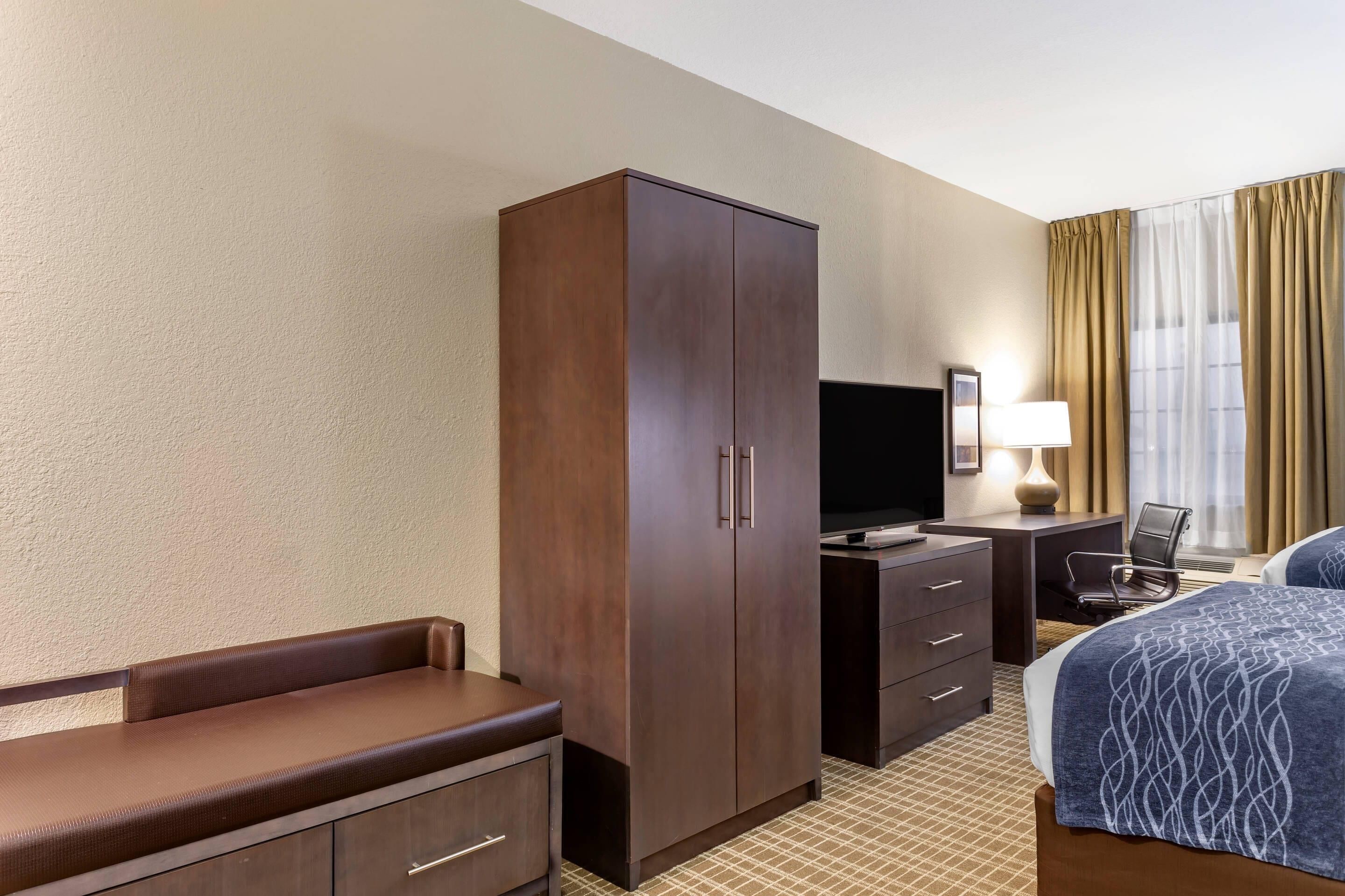 Habitación de hotel con un armario grande, un escritorio con lámpara, televisión, cama con colcha estampada y una silla.