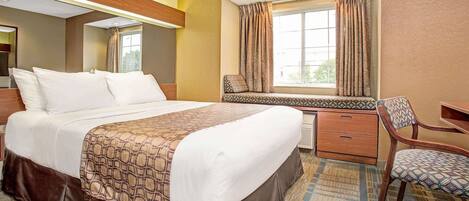 A hotel room with a large bed, a desk, a chair, a window with curtains, and a small seating area by the window.