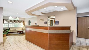 A reception area with a wooden reception desk, a window with curtains, a seating area with a chair and a table, and a door to the right. - Microtel Inn & Suites by Wyndham Mason/Kings Island (Mason)
