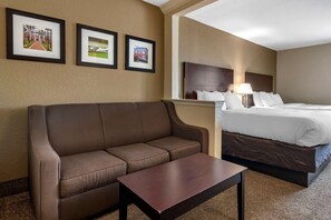 Une chambre d’hôtel avec un canapé marron, une table basse en bois, un lit avec du linge de lit blanc et trois tableaux encadrés accrochés au mur.