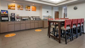 Een ontbijtruimte van een hotel met een lange balie, meerdere koffieautomaten en een zithoek met rode en witte stoelen.