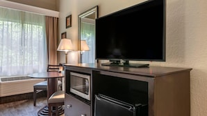 Een kleine hotelkamer met een flatscreen televisie, een magnetron, een koelkast, een eettafel met stoelen en een raam met dunne gordijnen.