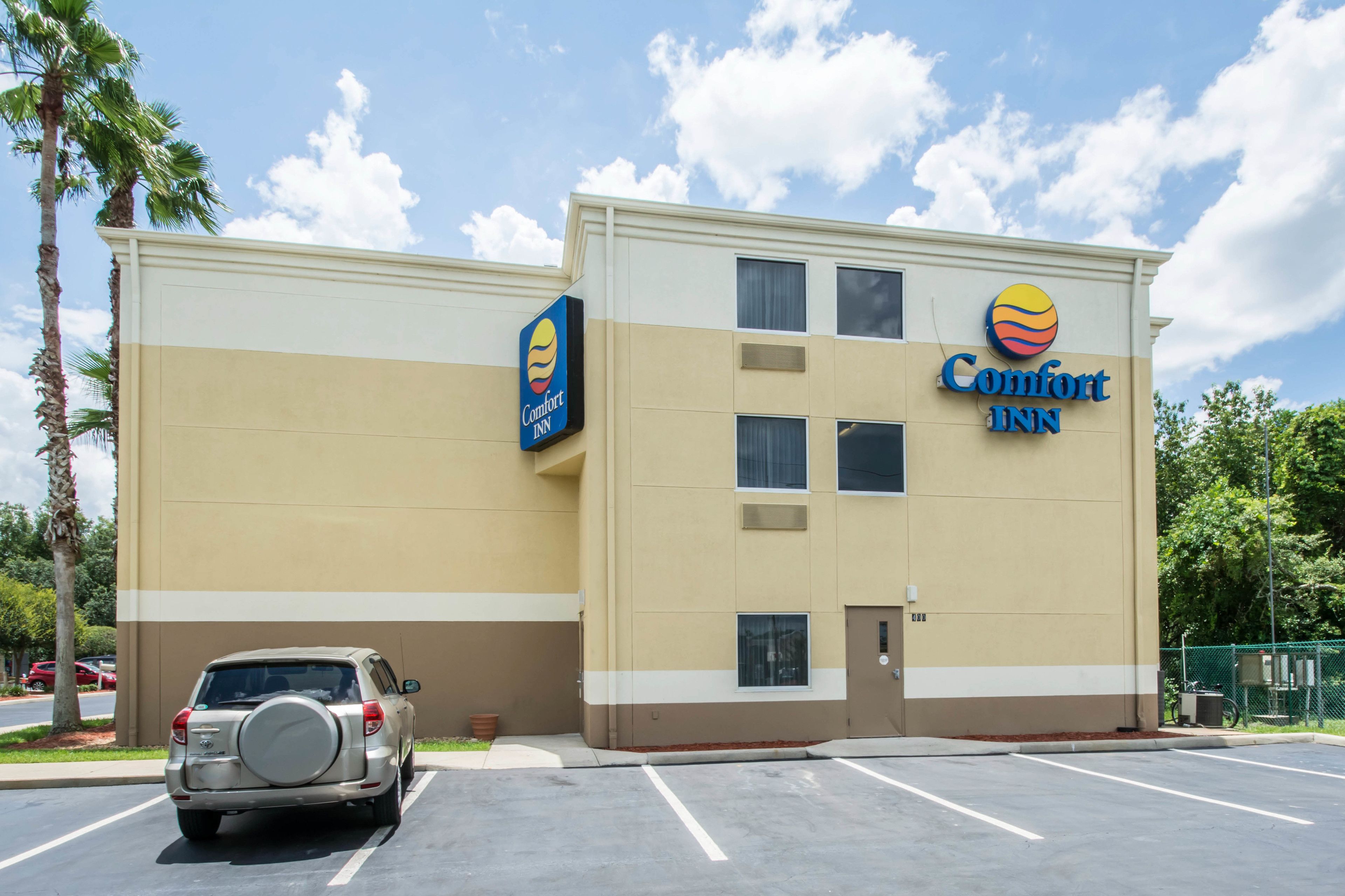 Um hotel Comfort Inn com fachada bege e branca, estacionamento e uma palmeira em primeiro plano.