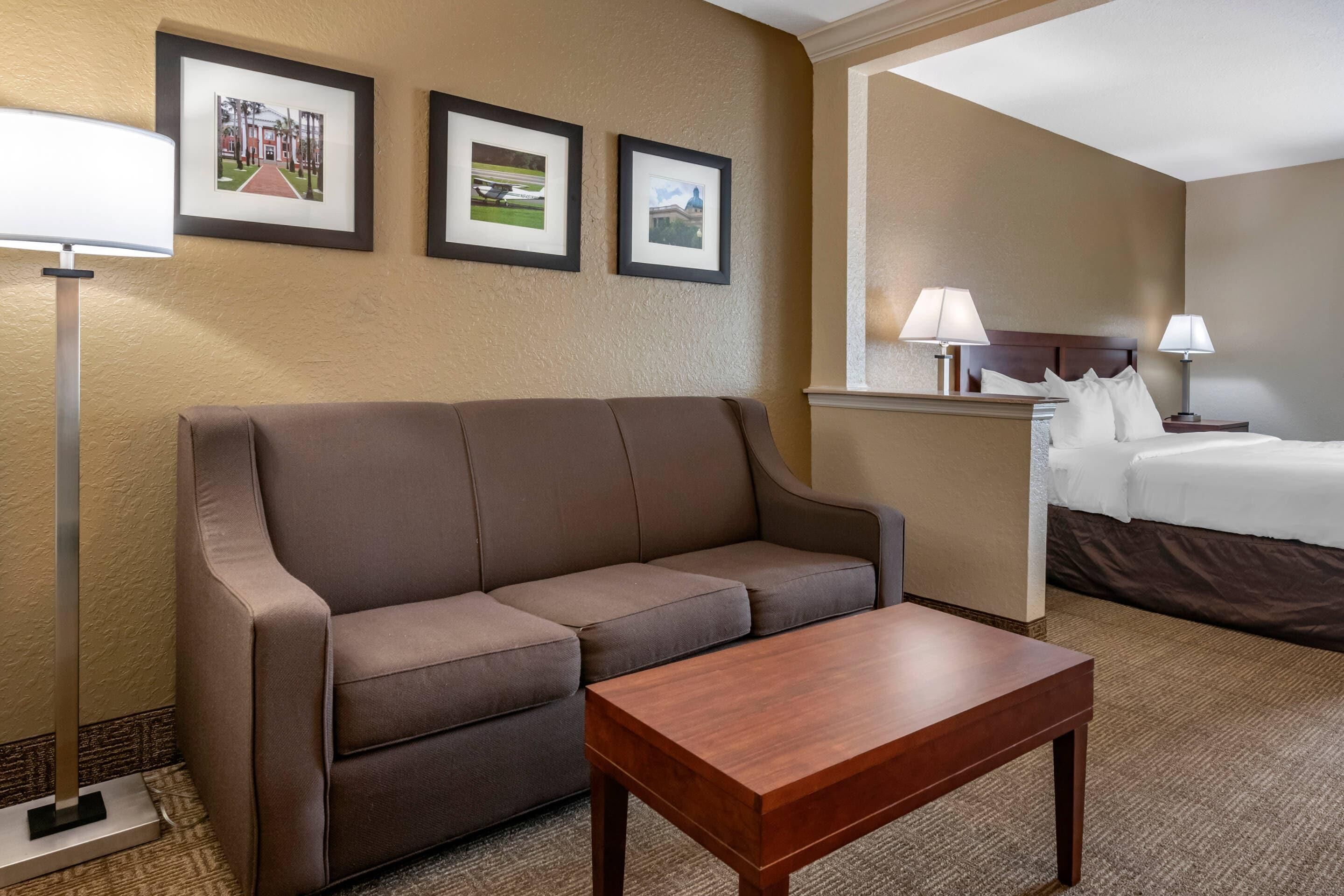 A hotel room with a brown sofa, a wooden coffee table, a bed with white linens, two bedside lamps, and three framed pictures on the wall.