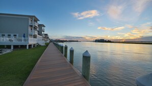 A wooden boardwalk leading to a waterfront property with a white building and a grassy area.