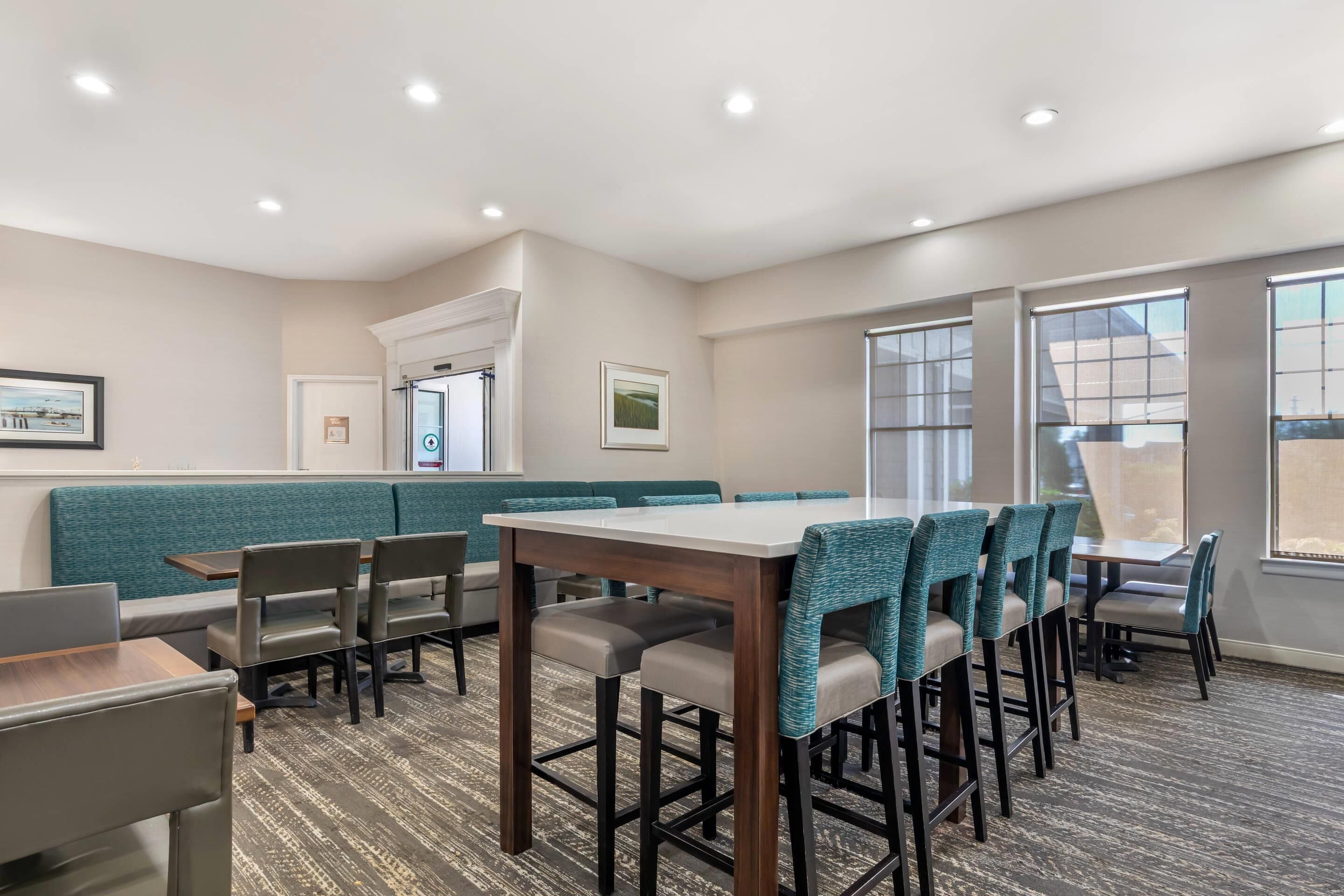 A hotel dining area with a long table, multiple chairs, a bench, and large windows.