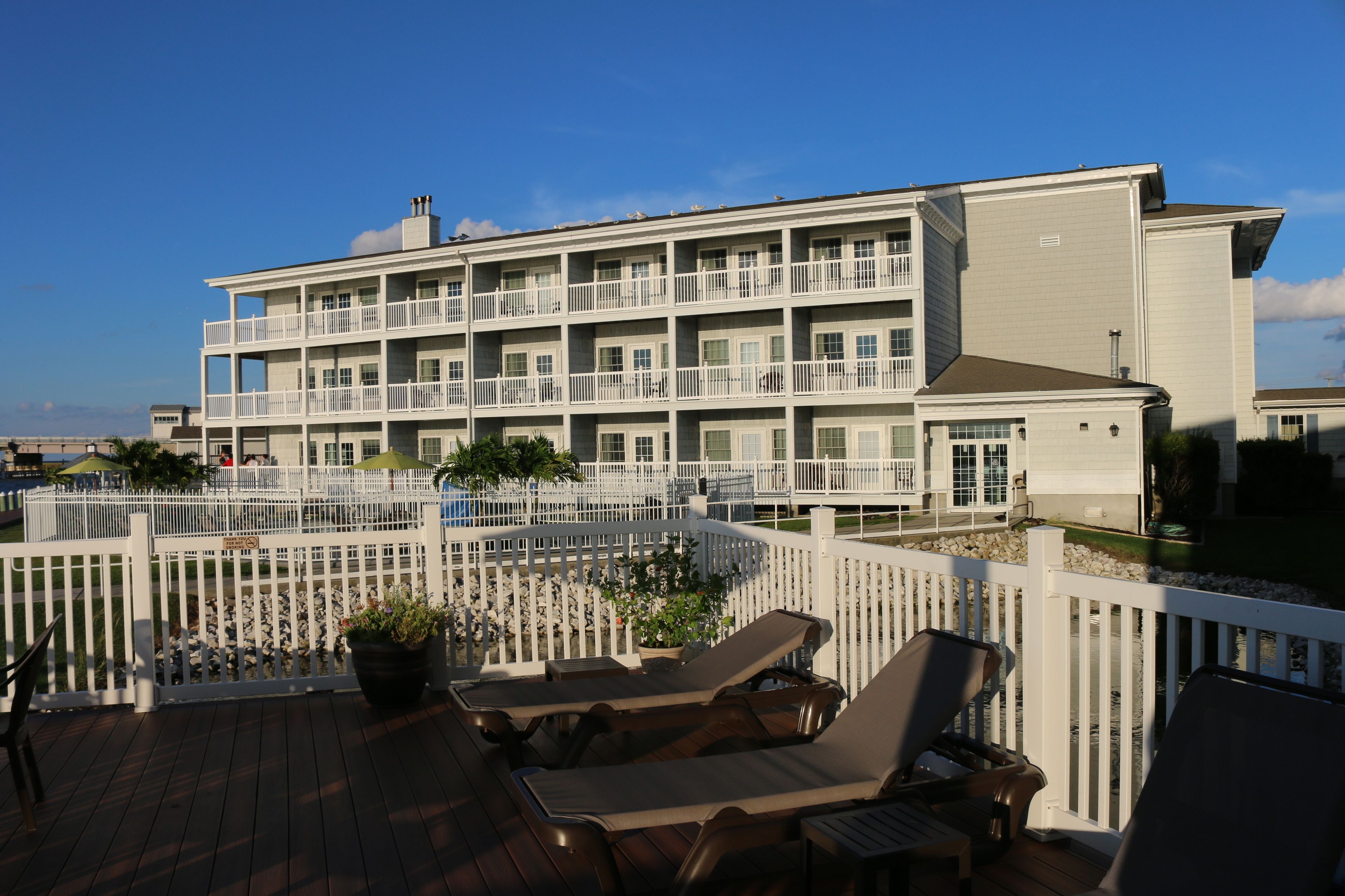 A multi-story hotel with balconies, a deck with lounge chairs, and a white fence.