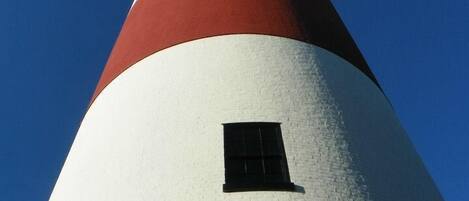Un faro alto y cónico con franjas rojas y blancas alternadas, una pequeña ventana y un cielo azul despejado al fondo.
