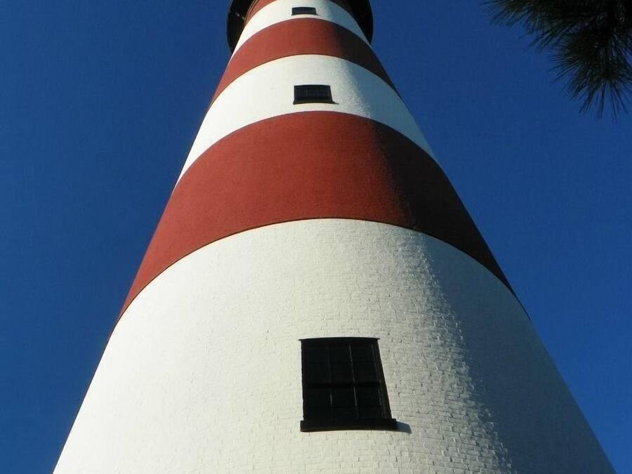 Un faro alto y cónico con franjas rojas y blancas alternadas, una pequeña ventana y un cielo azul despejado al fondo.