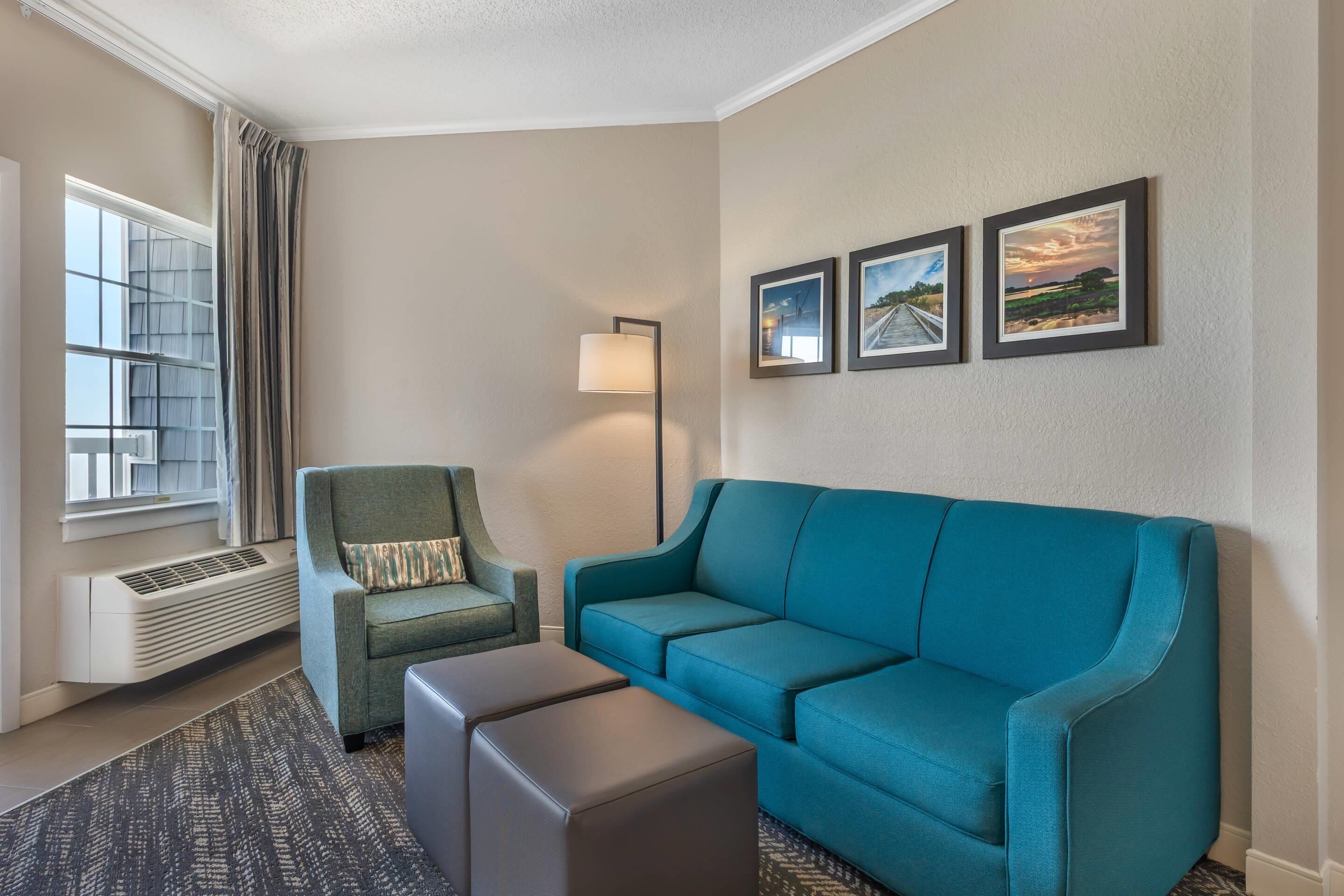A living room with a teal sofa, a green armchair, a floor lamp, and a radiator. There are three framed pictures on the wall.
