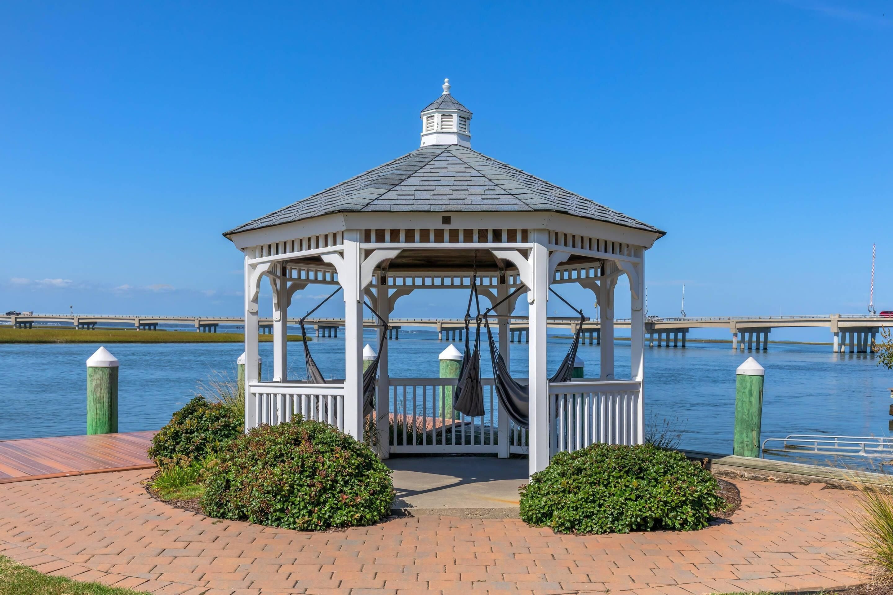 A gazebo with a white railing and a grey roof, situated by a body of water with a dock in the background.