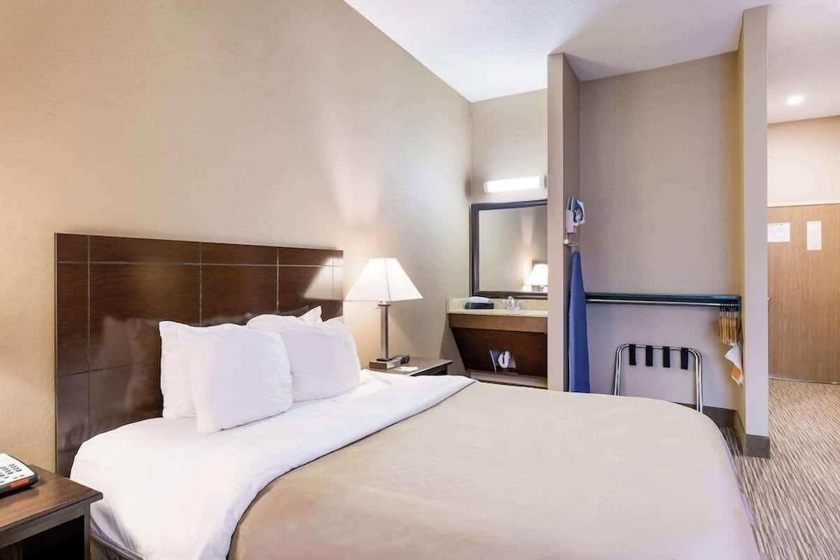 A hotel room with a large bed, a bedside table, a lamp, a mirror, a bathroom with a towel rack, and a door.