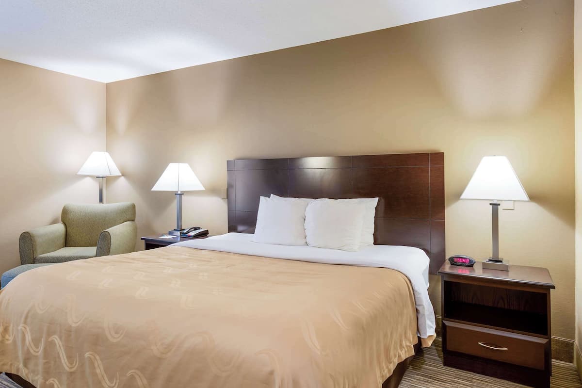 A hotel room with a large bed, two bedside tables with lamps, a chair, and a nightstand with a phone and a small electronic device.