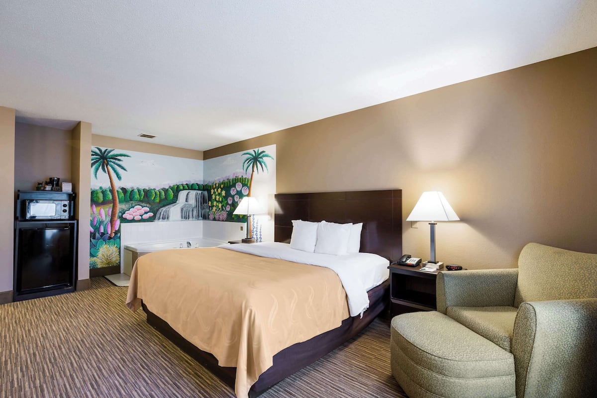 A hotel room with a large bed, a desk with a computer, a chair, and a painting of a waterfall and palm trees on the wall.
