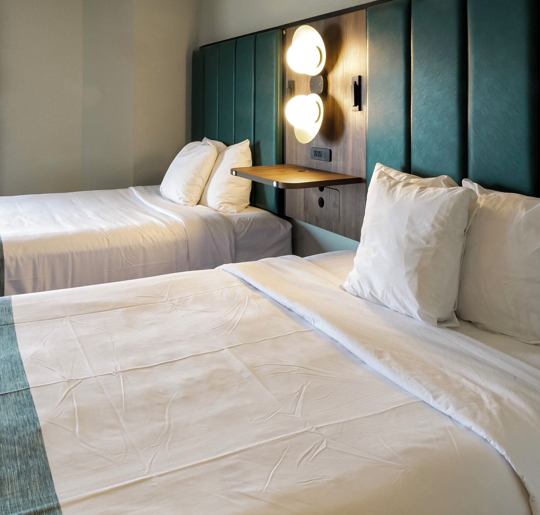 A hotel room with two beds, a nightstand with a lamp, and a wall-mounted light fixture.