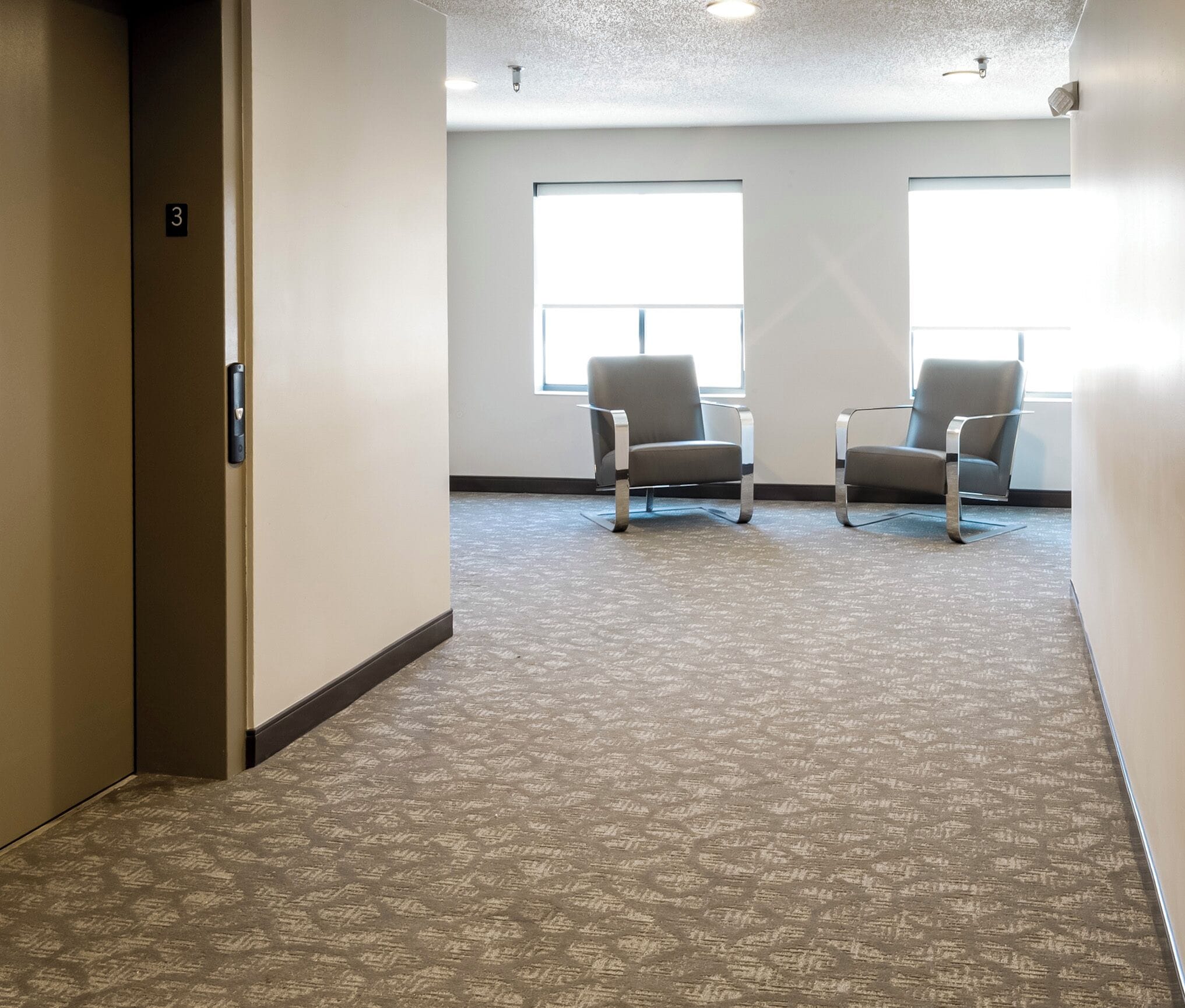 A hallway with carpeted flooring, two grey chairs, and a door marked with the number 3.