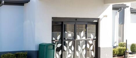 Entrada de un edificio con puerta de cristal, un bote de basura verde y un pequeño jardín.