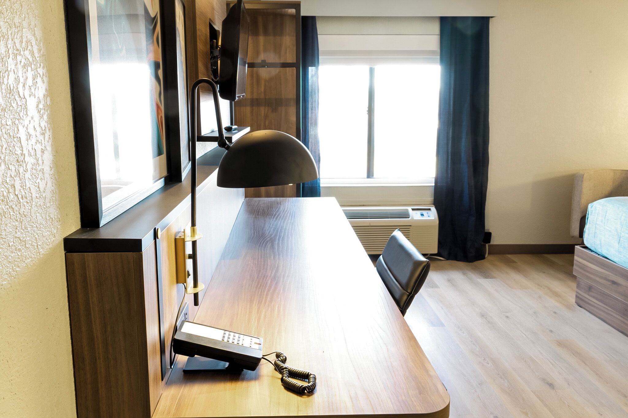 Una habitación de hotel con un escritorio de madera, un teléfono, una lámpara, una cama y una ventana con cortinas.