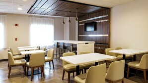 A modern dining area with multiple tables and chairs, a kitchen area with a flat-screen TV, and a large window with curtains. - Wingate by Wyndham Owensboro (Owensboro)