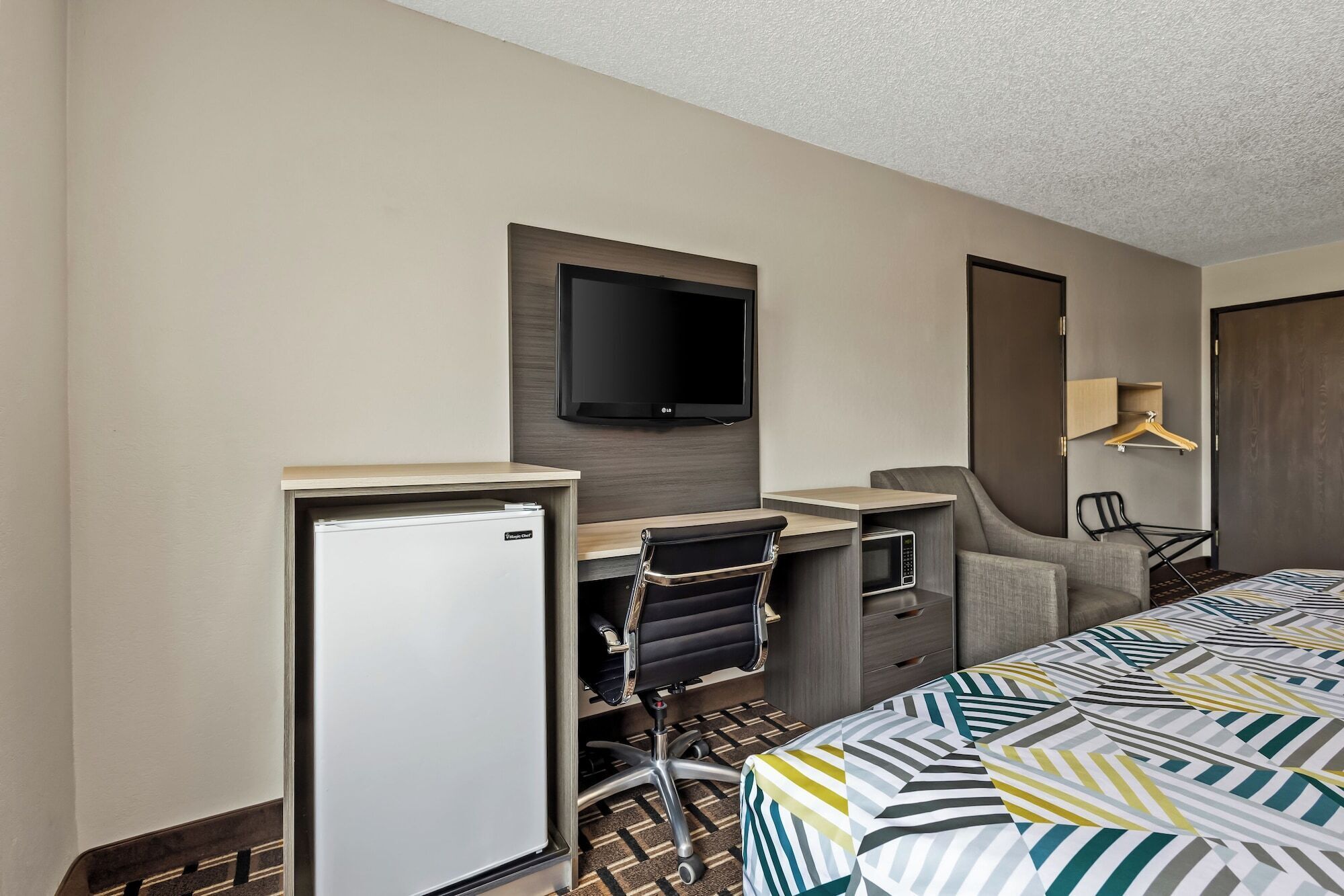 A hotel room with a bed, a desk with a chair, a television, a refrigerator, and a wardrobe.