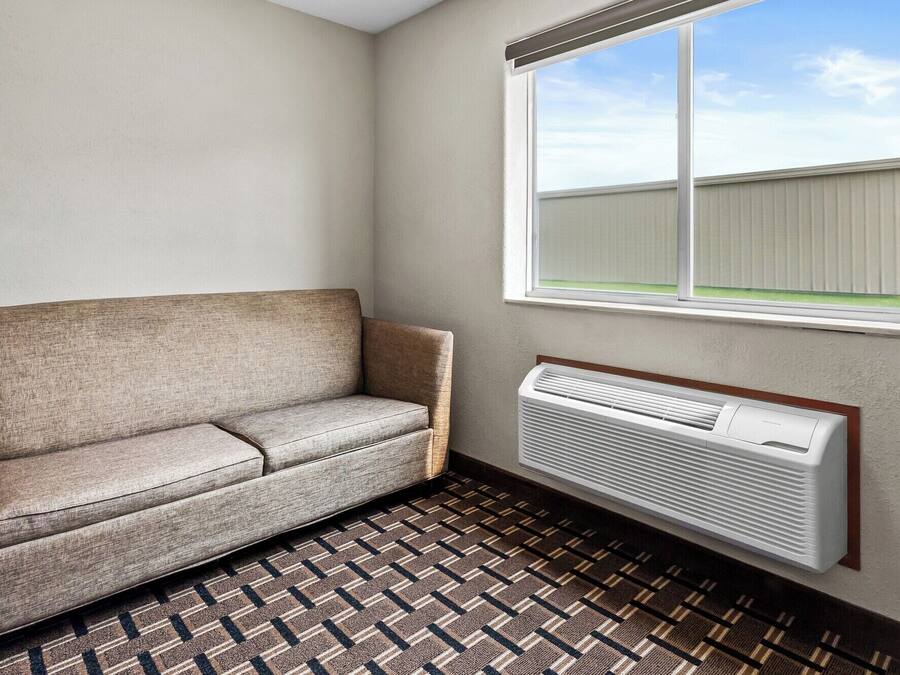 Una habitación con sofá color beige, alfombra estampada, ventana con vista a un edificio y aire acondicionado instalado en la pared.