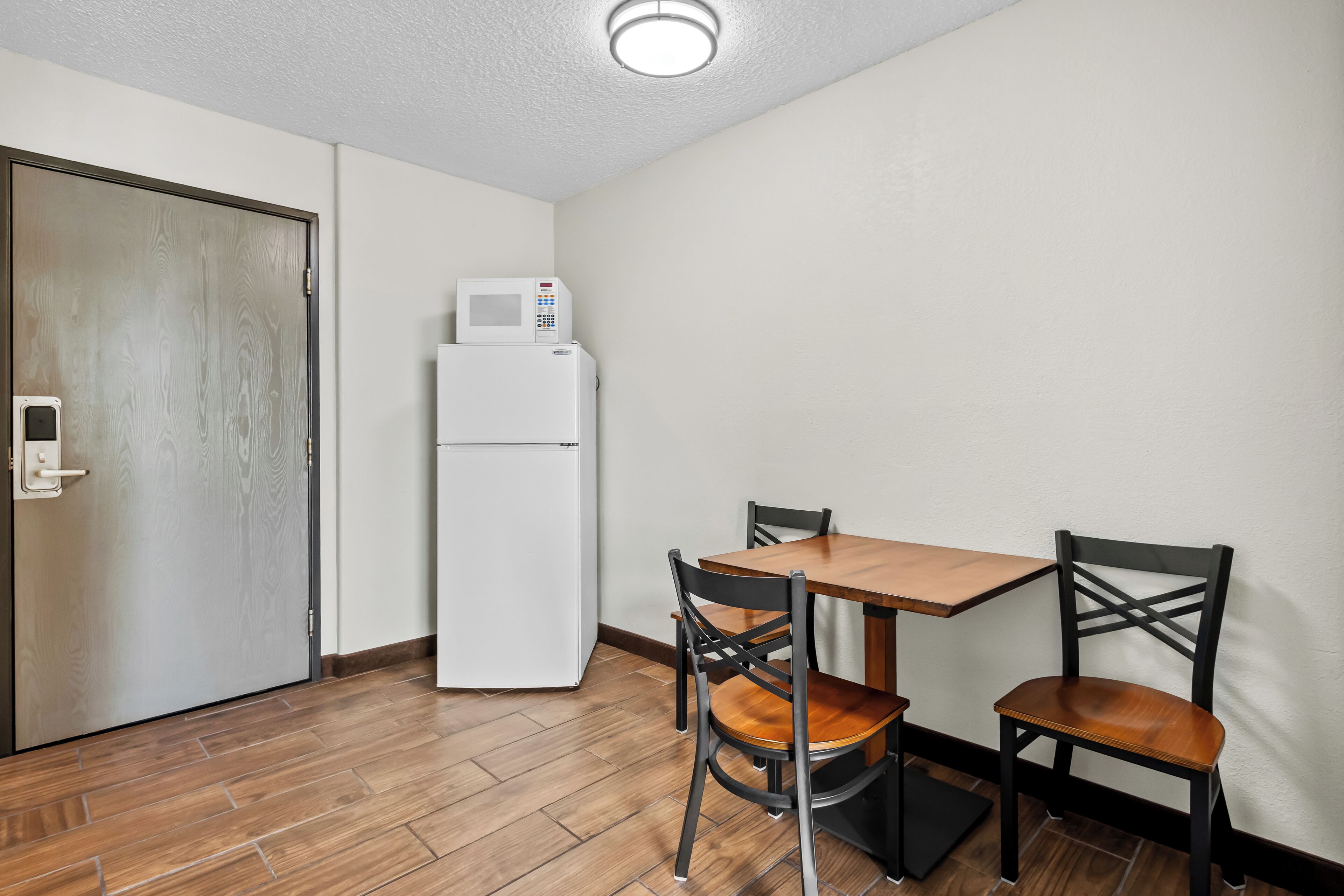 Una pequeña cocina americana equipada con microondas y refrigerador, una mesa de comedor de madera con dos sillas, y una puerta con picaporte.