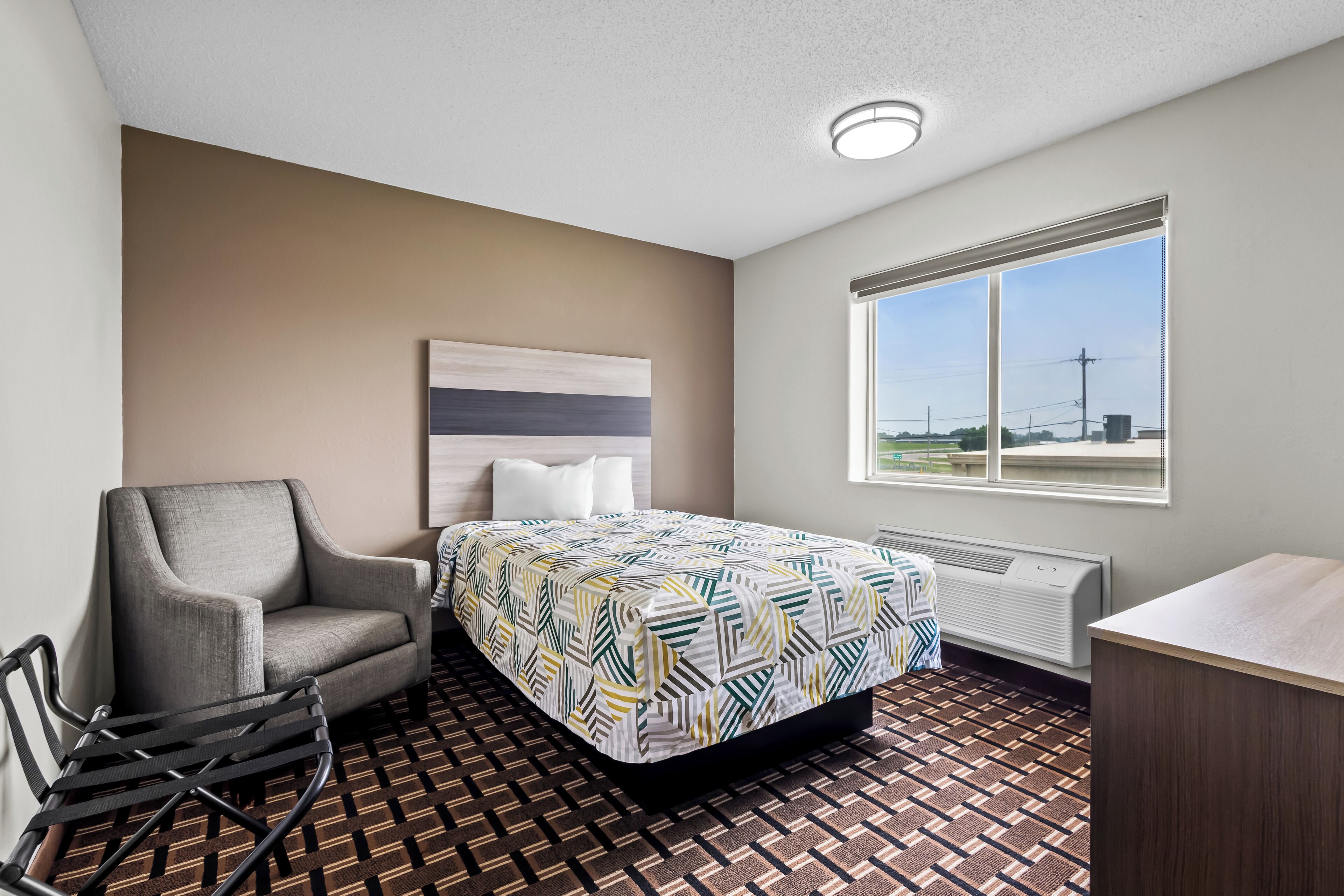 A hotel room with a bed, a chair, a desk, a radiator, and a window with a view.