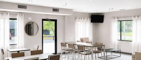 A hotel dining area with multiple tables and chairs, a flat-screen TV mounted on the wall, and large windows with curtains.