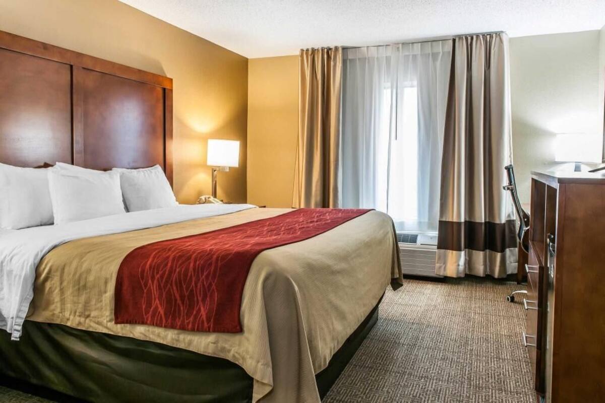 A hotel room with a bed, a desk, a lamp, a window with curtains, and an air conditioning unit.