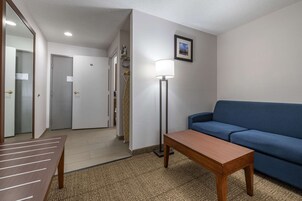 A hotel room with a blue sofa, a wooden table, a floor lamp, and a door leading to a hallway.