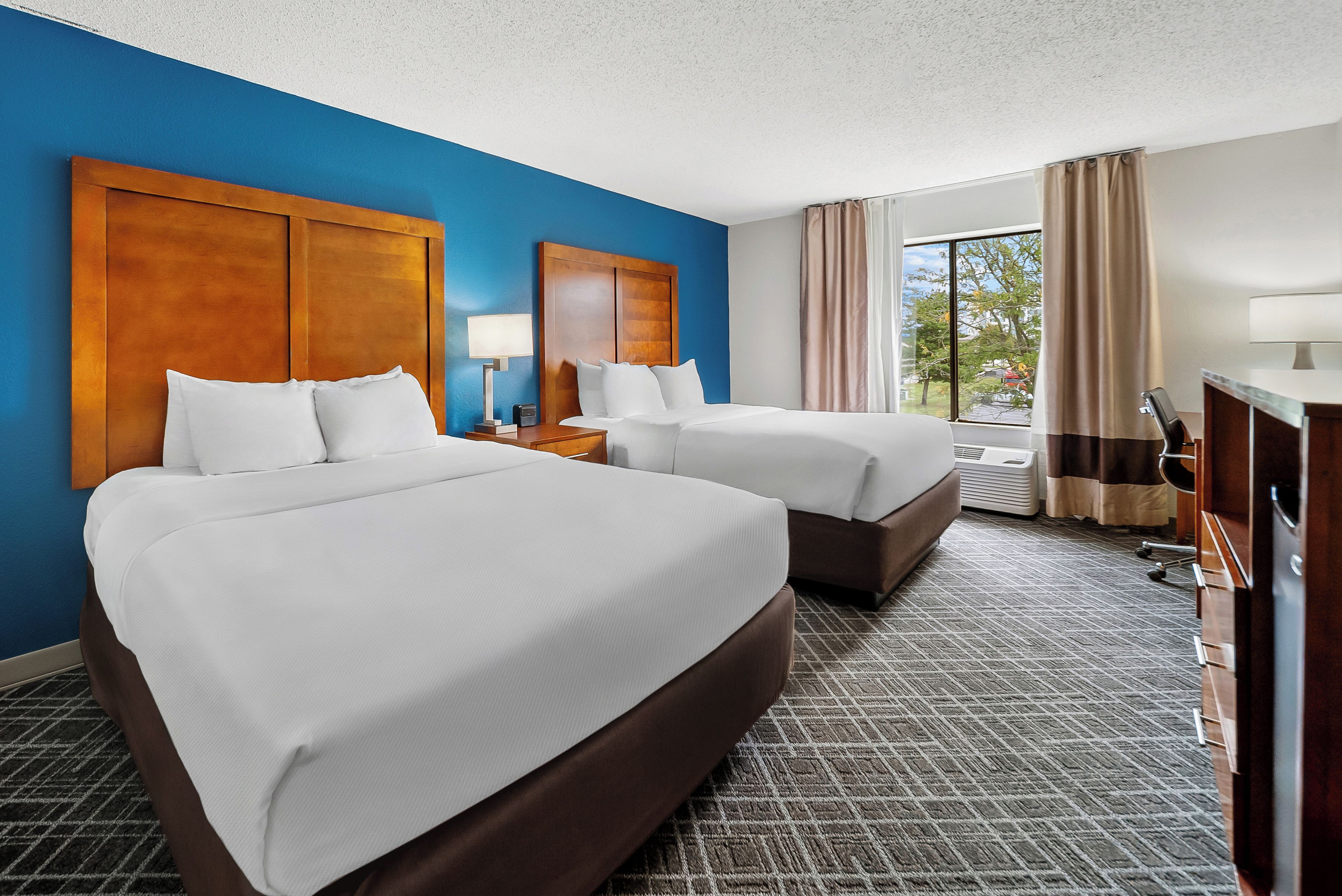 A hotel room with two beds, a desk, a lamp, a window with curtains, and a view of the outdoors.