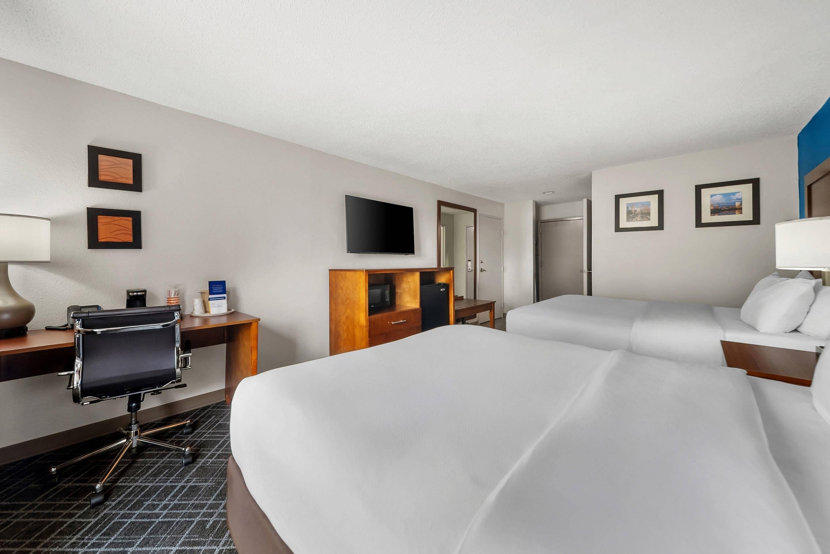 A hotel room with two beds, a desk with a chair, a flat-screen TV, a minibar, and a door leading to another room.
