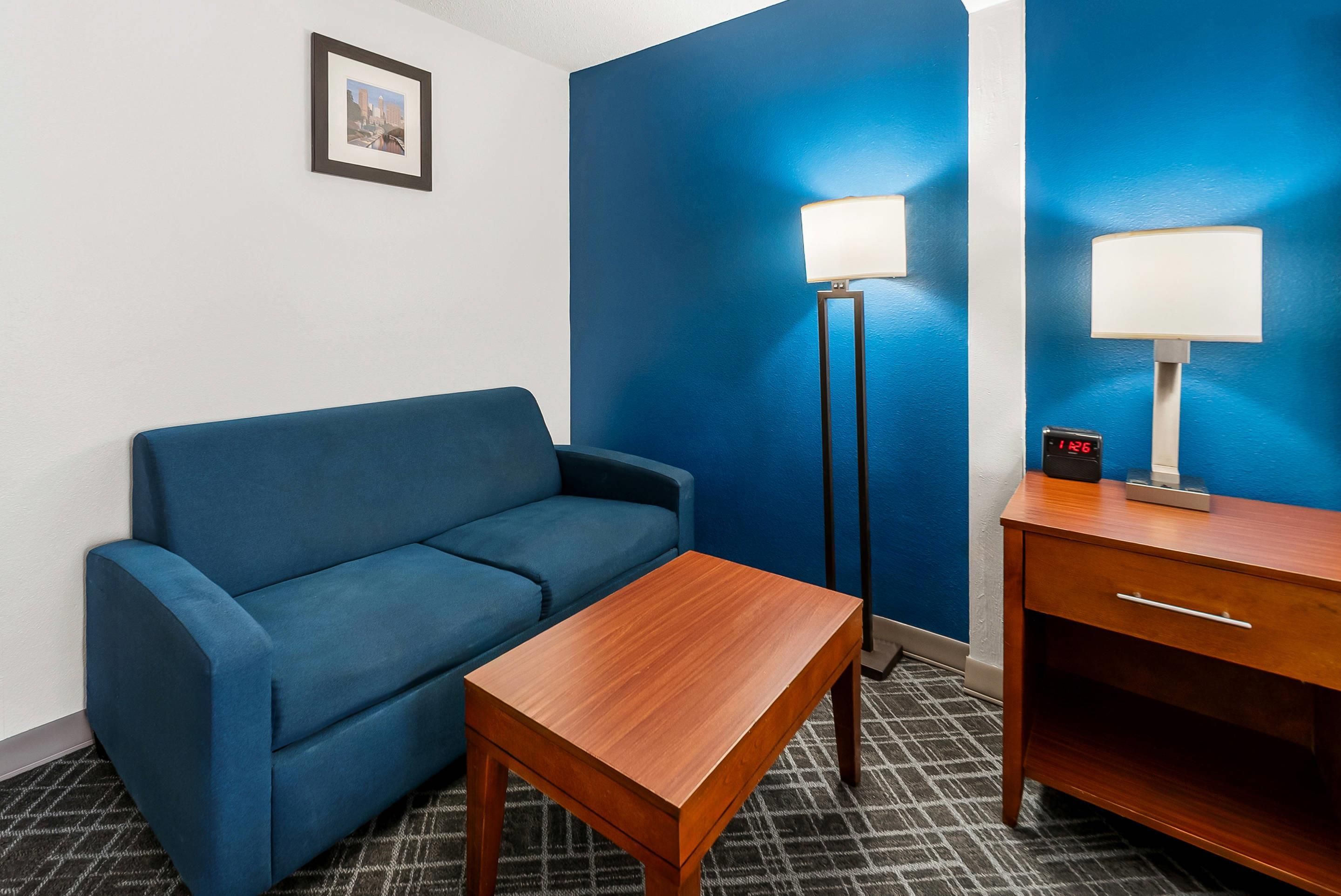 Une chambre d’hôtel avec un canapé bleu, une table en bois, une lampe sur pied, une table de chevet avec une lampe et un tableau encadré au mur.
