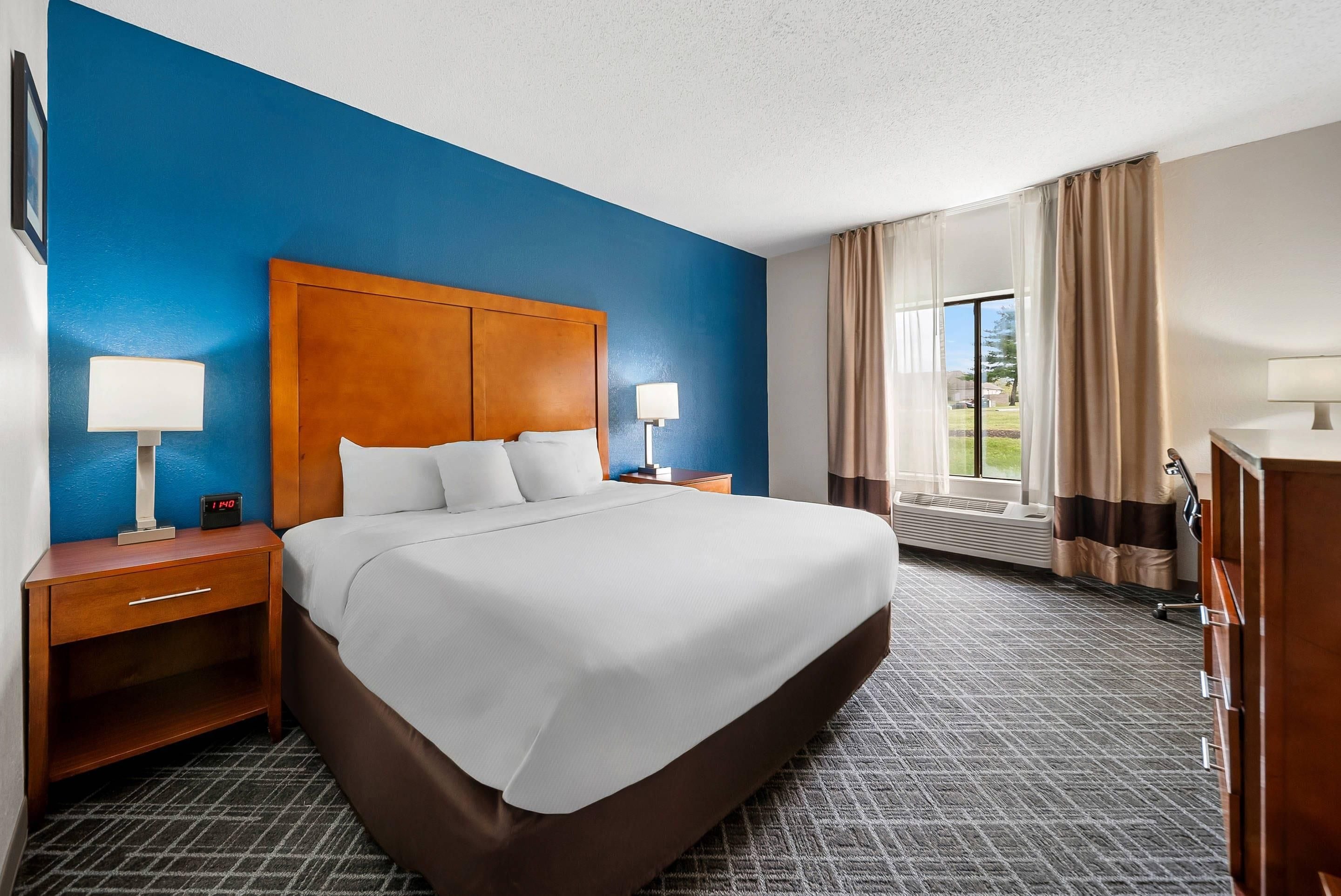Una habitación de hotel con una cama grande, dos mesitas de noche con lámparas, una mesita auxiliar, un aparador y un ventanal con cortinas.