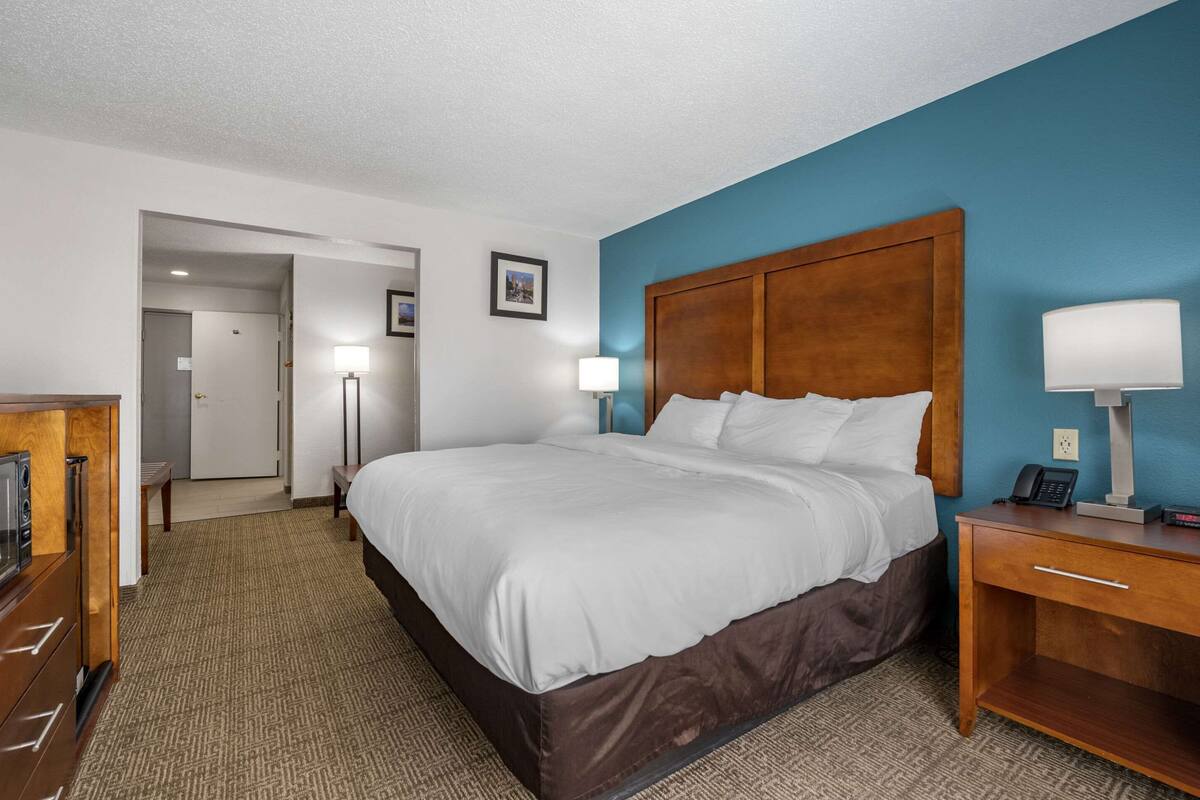 A hotel room with a large bed, a wooden nightstand with a lamp, a desk with a phone, and a view of the hallway.