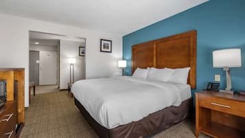 A hotel room with a large bed, a wooden nightstand with a lamp, a desk with a phone, and a view of the hallway.