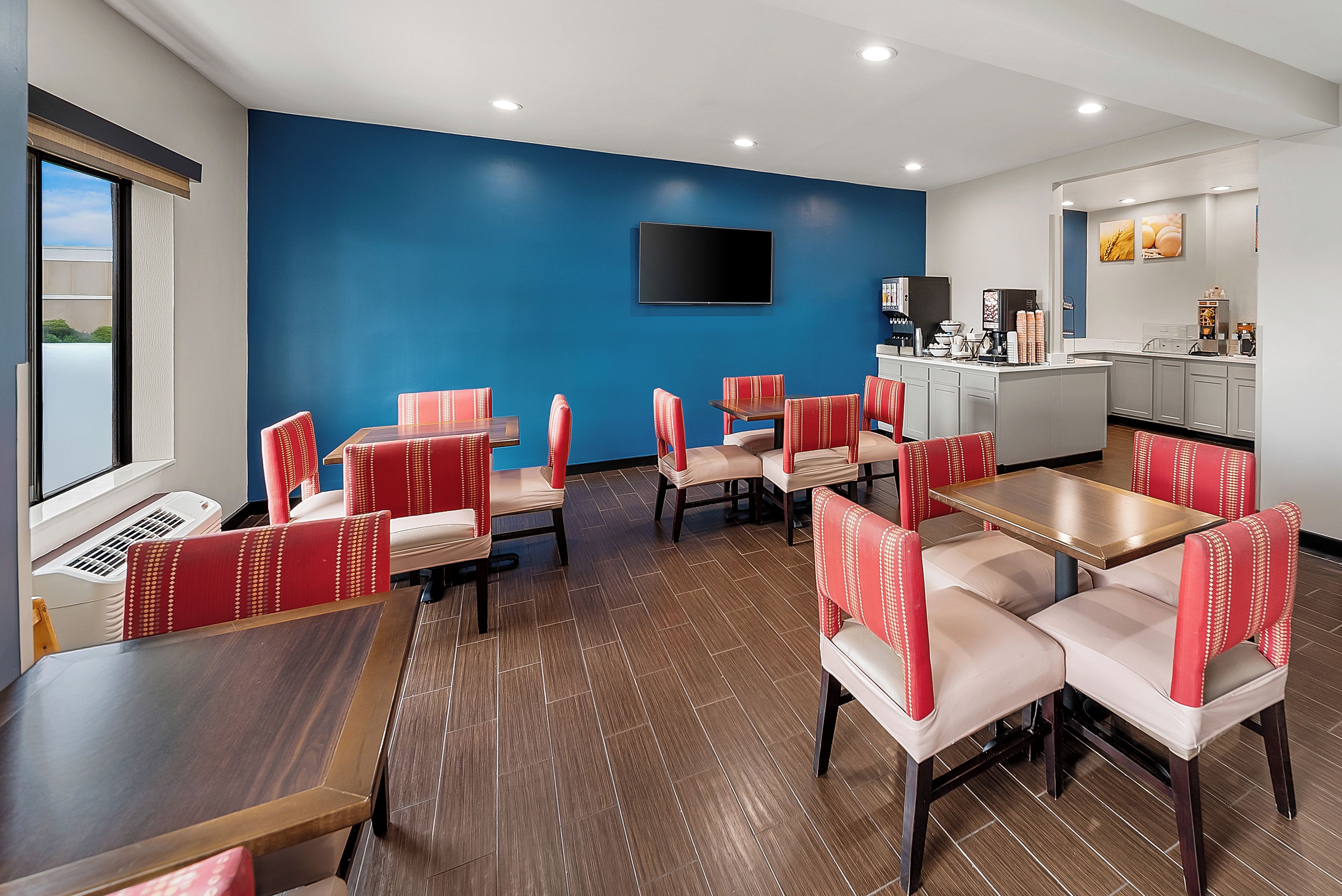 Un comedor de hotel con asientos tipo banca tapizados en rojo y beige, un televisor de pantalla plana en la pared y una kitchenette con máquinas de café.